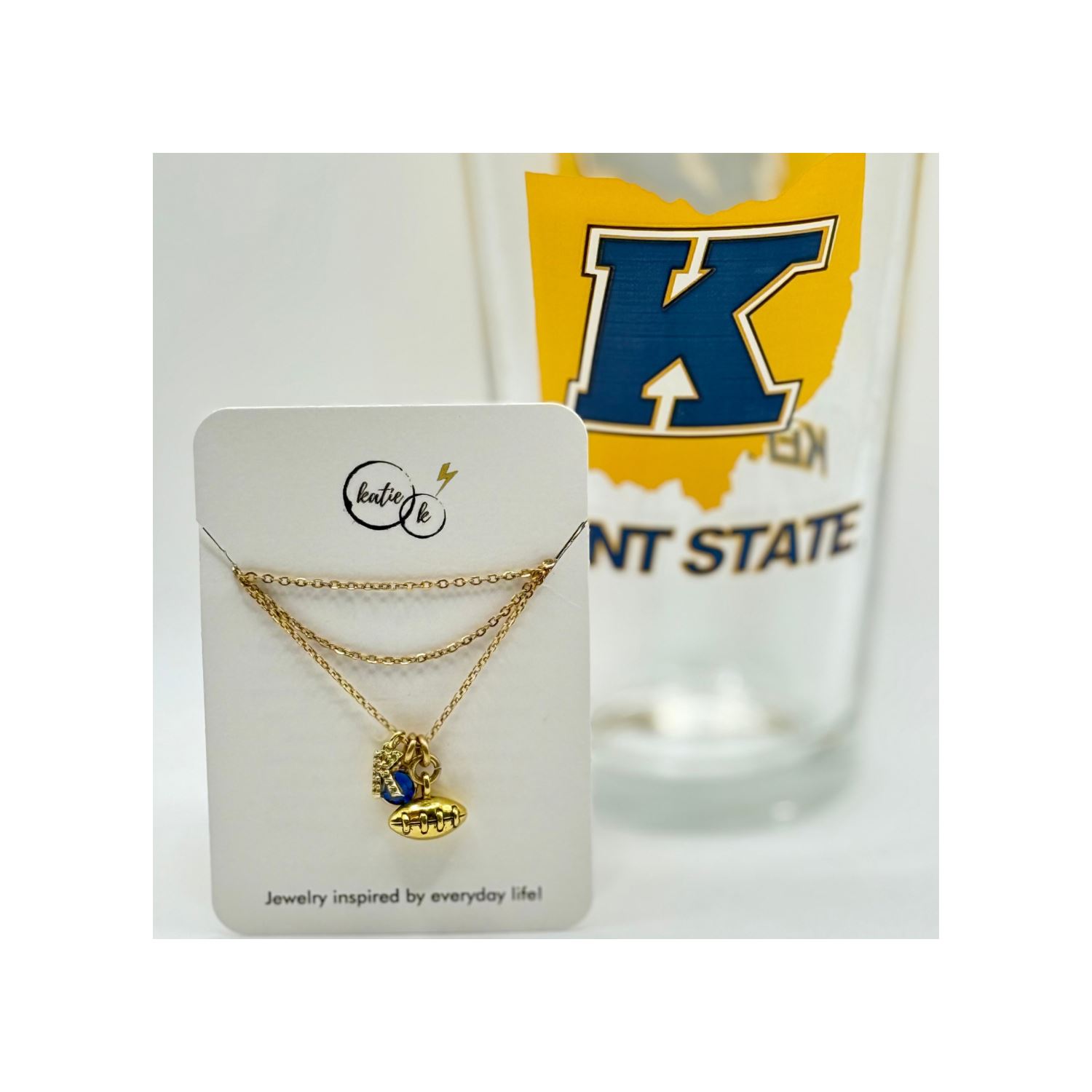 Mini Sparkly K Sapphire Crystal And Textured Football Charm Necklace displayed on a card with a Kent State University themed glass in the background.