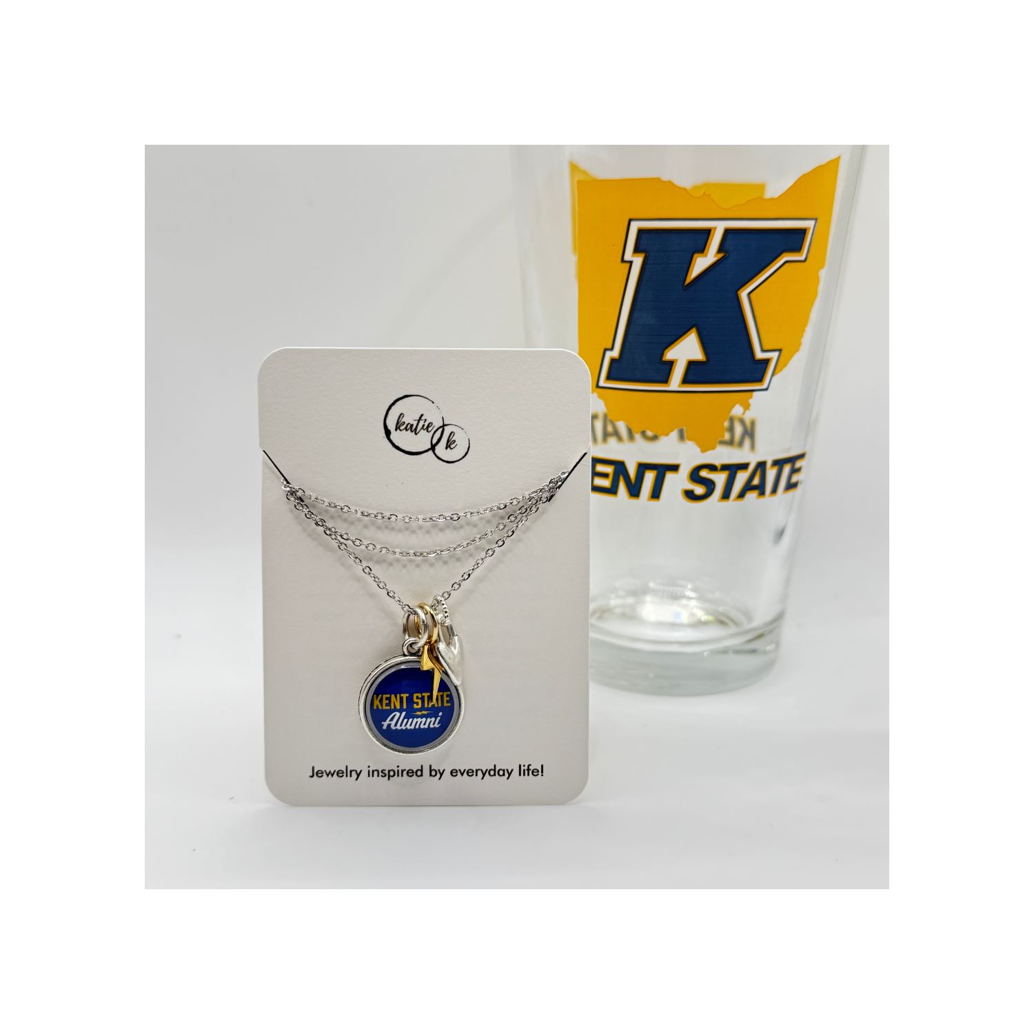 Kent State Alumni Charm Necklace featuring a silver chain with a blue and gold round charm and a small silver key charm, displayed with a Kent State University glass in the background.